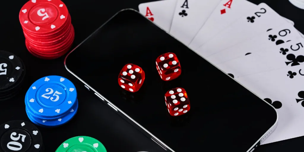 Foto ilustrativa com fichas de cassino, cartas de baralho e dados vermelhos sobre um celular escuro.